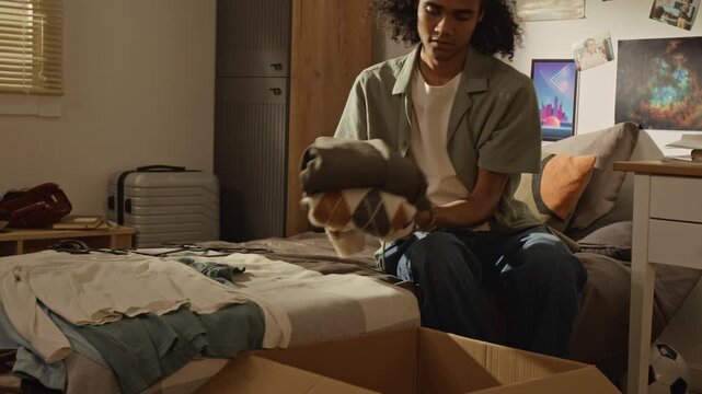 Handheld shot of Black male teenager folding clothes and packing cardboard box in bedroom, preparing for relocation. Footage is ideal for moving services, student housing, or decluttering concepts