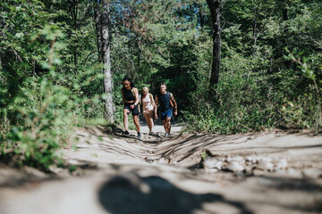 Obraz premium Three friends hike up a sandy forest trail together. The group walks through green woodland on a sunny day for exercise.