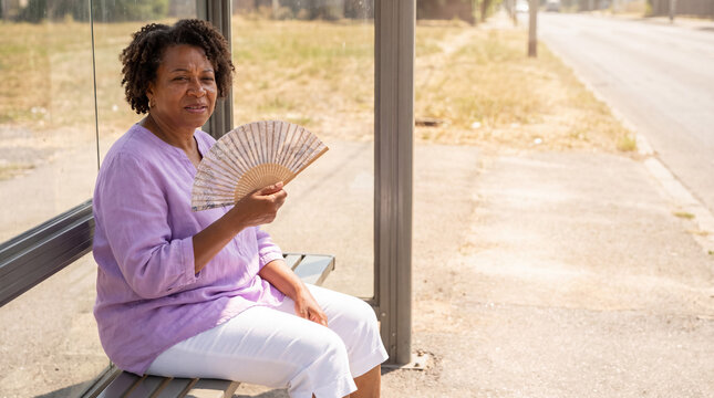 Mature woman cooling with hand fan, experiencing hot flash while waiting at a sunny bus stop