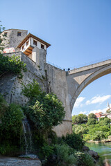 Fototapeta premium MOSTAR, BOSNIA AND HERZEGOVINA Low-angle vertical view of the historic Halebija Tower and Stari Most stone arch, featuring a natural small waterfall cascading down green bushes.
