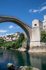 Naklejka premium Low-angle view of the iconic Stari Most (Old Bridge) stone arch and the historic Tara Tower overlooking the green Neretva riverbank MOSTAR, BOSNIA AND HERZEGOVINA