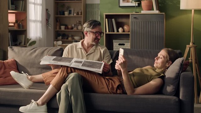 Wide shot of middle aged Caucasian couple relaxing on sofa, man reading newspaper while woman browses smartphone. Clip ideal for advertising digital media transition or home leisure products