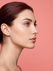 Fototapeta premium Elegant studio portrait of a young woman in side profile featuring flawless glowing skin, soft natural makeup, and a warm pink background with premium beauty styling.