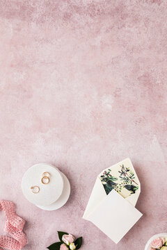 Studio shot of invitation card, ribbon, engagement ring and pair of wedding rings flat laid against pink background
