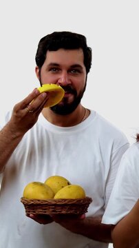 Indian young couple eating mango while standing isolated against white background, holding, promoting fresh seasonal aam or Alphonso happily, enjoying king of fruits with joyful expressions smiles