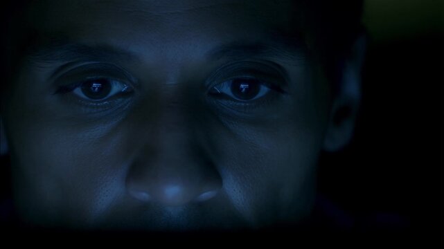 A man's face is illuminated by a blue screen in the dark. He stares intently with wide eyes, blinking occasionally, capturing a moment of deep focus or digital fatigue.
