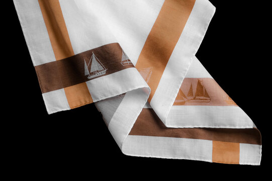 A close-up shot of a white cotton men's handkerchief decorated with brown stripes and a repeating sailboat pattern against a black background.
