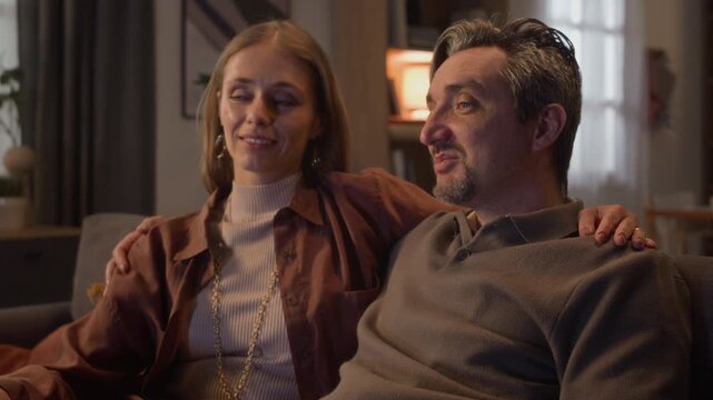 Close up of middle-aged Caucasian couple sitting on sofa, man using remote control to switch channels before talking and smiling. Video suitable for smart TV, streaming service or cable provider ads