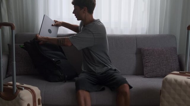 Always working lifestyle and nonstop mobility. Man working on laptop on sofa with luggage, finishing work and leaving apartment immediately