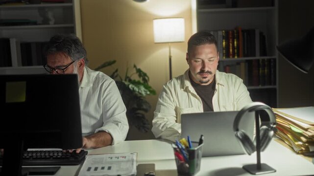 Two men at computers, one hand to chin over a laptop beside desktop monitor and stacked folders in a building; quiet concentration.