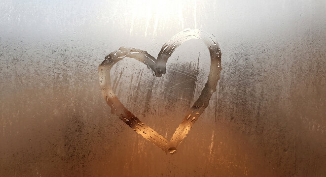 Heart shape drawn by finger on a foggy window with water drops and warm glowing light