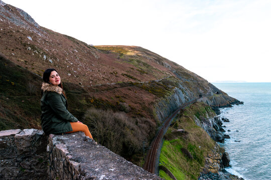 Young woman sitting on a stone wall overlooking scenic coastal railway and cliffs in Ireland