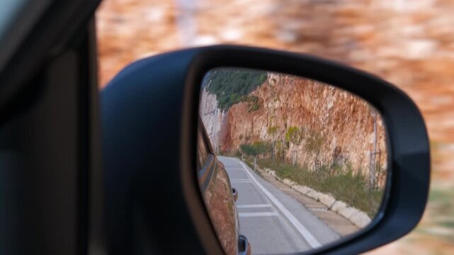 Car side mirror reflecting winding countryside road. Rear-view mirror reflecting a scenic mountain road during a road trip through a rocky landscape, showing the journey's progress with a blurred