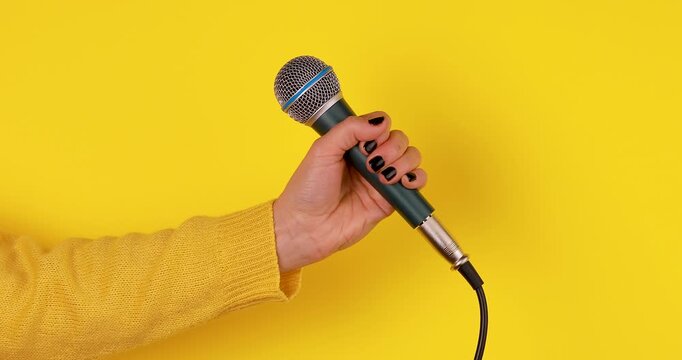 Close-up video of a female hand holding a microphone against a yellow background and pointing it toward herself. Concept of speaking, self-expression, interviewing, podcasting, media, and communicatio