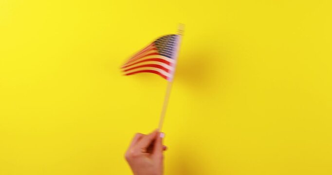 Close-up video of a hand waving the American flag against a bright yellow background. Patriotic concept symbolizing freedom, national pride, celebration, identity, and United States culture.