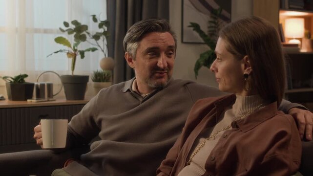 Medium shot of middle aged Caucasian couple sitting on sofa, talking and embracing while woman kisses man on cheek. Footage for relationship counseling, family insurance, or home lifestyle commercials