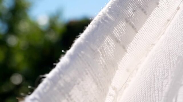 Sun-kissed white linen fabric waving gently in the breeze. Macro detail emphasizes the crisscross pattern of the weave and the natural, airy quality of the material. Bright daylight.
