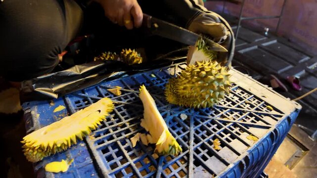 Close-up of a Thai street vendor cutting open a fresh durian with a large knife while wearing a protective glove at a local night market. Authentic tropical fruit preparation captured in 4K.