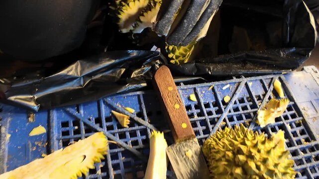 Close-up of a Thai street vendor slicing through a ripe durian with a large knife while wearing a protective glove at a local night market. Authentic tropical fruit preparation in Thailand, captured i