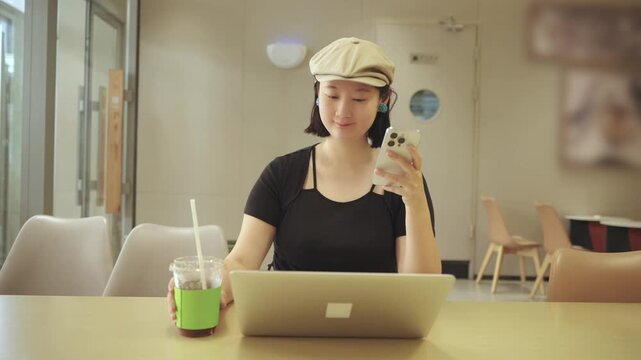 A young asian woman uses a smartphone while sitting at an indoor table with a laptop and a drink, in a casual setting.