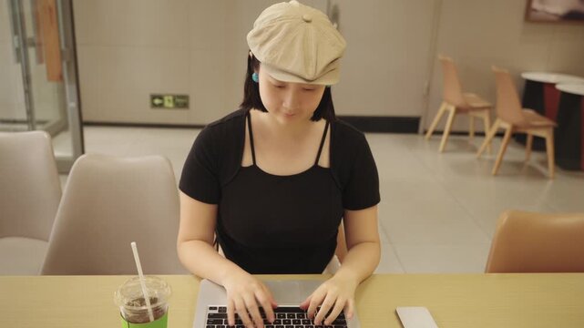 A young woman wearing a beret uses a laptop while seated at a table in an indoor cafe, with a drink placed beside her.