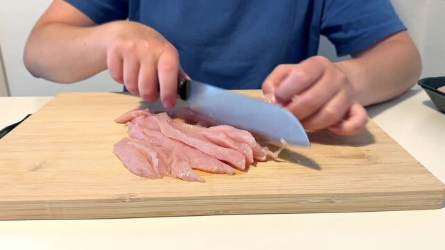 Cutting raw chicken breast on wooden cutting board with sharp knife in kitchen at daytime for food preparation