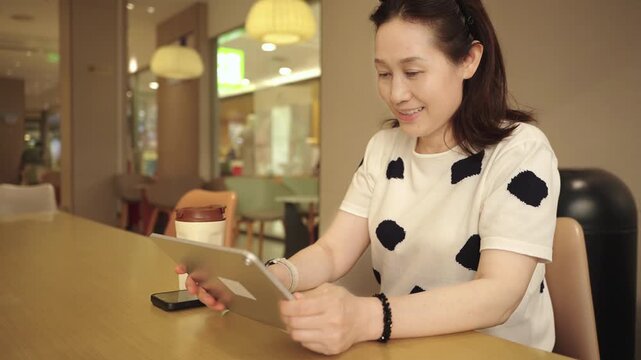 A smiling middle-aged asian woman uses a tablet while sitting at a wooden table in a cozy cafe interior. A drink cup and smartphone are also on the table.