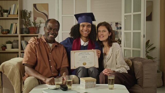 Medium long portrait of proud middle aged Black father, Caucasian mother and teenage son in graduation cap holding certificate at home. Ideal for education marketing and family success campaigns