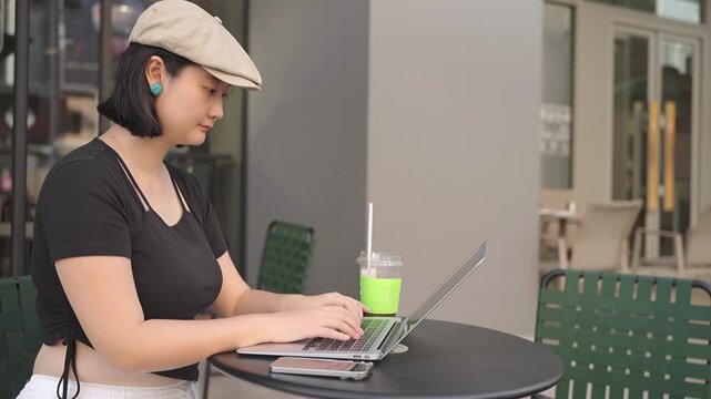 An Asian adult woman sits at an outdoor cafe table, using a laptop with a drink and smartphone nearby. She wears casual clothing and a hat.