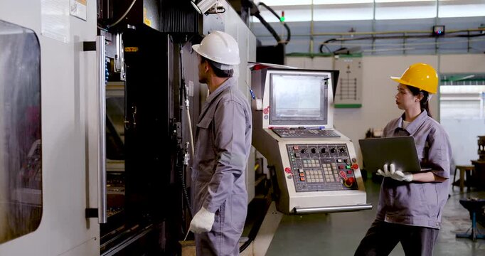 Asian man engineer checks CNC machine area as Asian woman technician reviews laptop data, coordinating machine status, process check, and production tracking for factory workflow, slow motion