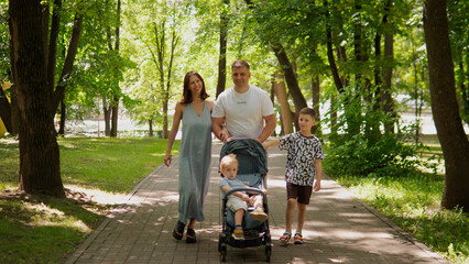 Naklejka premium Father and mother stroll along tree-lined paved park path with toddler seated in stroller and older child walking beside them; dappled sunlight filters
