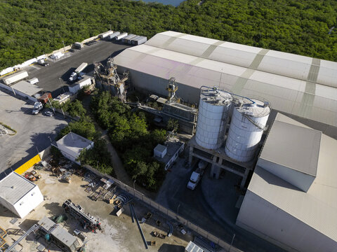 Aerial view of Ergon Asfaltos Progreso industrial facility with storage silos, warehouse buildings, and trucks in the yard, surrounded by dense vegetation in Progreso, Yucatan, Mexico.