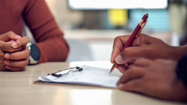 Two people sit at a table, one signing a document. Hands steady, focused, suggesting agreement or commitment. Clipped papers and a red pen draw attention to the action. The setting feels professional