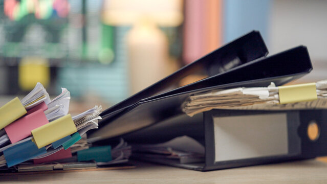 Stacked binders overflow with papers, showing chaotic work. Cluttered desk hints at urgent deadlines or overwhelmed tasks. Soft-focus background suggests busy office or study space
