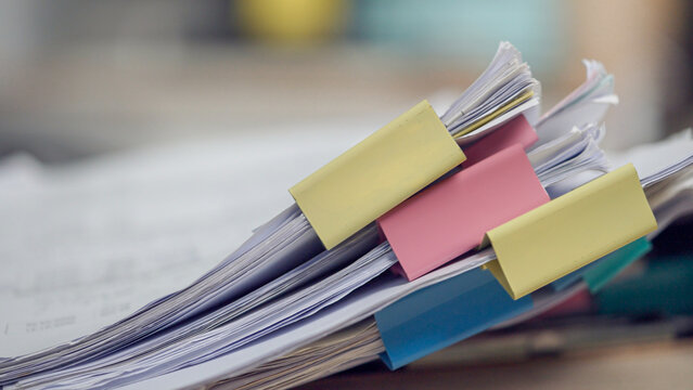 Stacked papers binded with colorful clips. Blurry background suggests busy office space. No people are visible in the frame. Ideal for business, organization, or paperwork themes