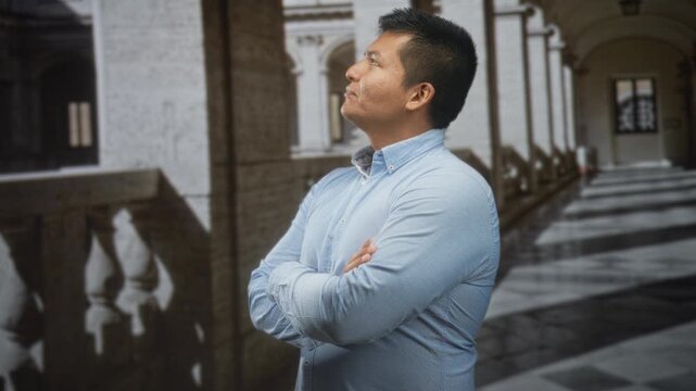 Man with arms crossed by a stone railing in a colonnaded building, wearing a blue buttondown shirt and looking upward; quiet contemplation.