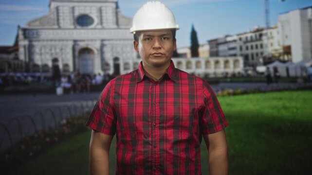 Man engineer wearing hardhat and red plaid shirt stands facing camera with forearms visible in front of ornate cathedral facade at building plaza; duty.