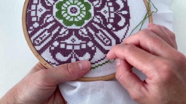 Close-up of a wooden hoop with traditional cross-stitch. A woman embroiders a purple-green floral motif, decorative ornament, creating a beautiful painting