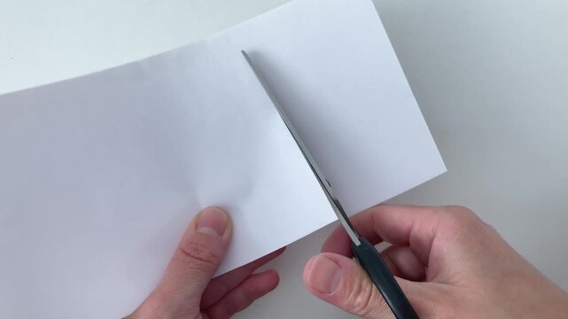 Top view of a woman's hands with scissors on a white background. A woman cuts a sheet of white paper. Preparation of materials for stationery and decorative products, creativity