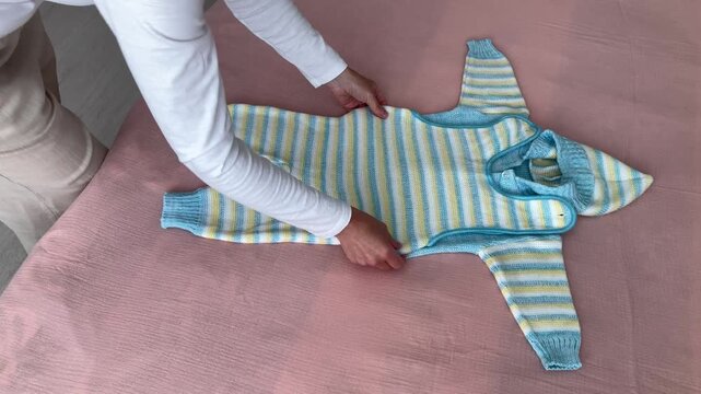 A woman carefully lays out a knitted baby jumpsuit on the bed, preparing things for the birth of a baby. Warm clothes for the cold season. Side view