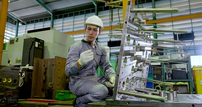 Asian technician kneels beside large metal fixture while holding guide rod, checking alignment, production readiness, safety step, precision setup, maintenance workflow in , slow motion.
