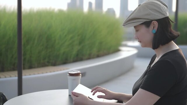 An asian young woman uses a tablet while seated at an outdoor urban patio, with a coffee cup beside her.