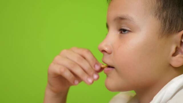Close up of a boy eating a salted breadstick snack, taking a crunchy bite with a green background indoors.