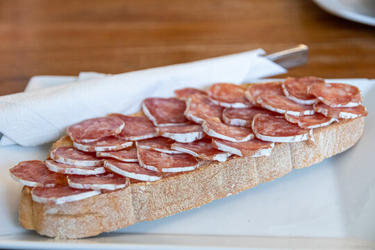 Vielha, Spain. Close up of a traditional breakfast toast topped with slices of cured fuet sausage and extra virgin olive oil, served on a white plate with a napkin inside a local restaurant