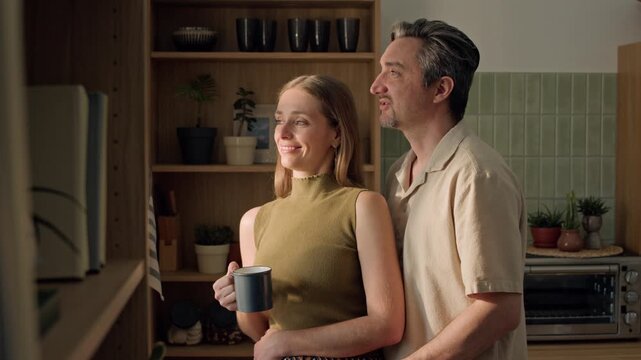 Medium shot of middle aged Caucasian man and young adult Caucasian woman standing in kitchen, looking out window, holding mug and kissing. For real estate, home insurance or coffee brand advertising