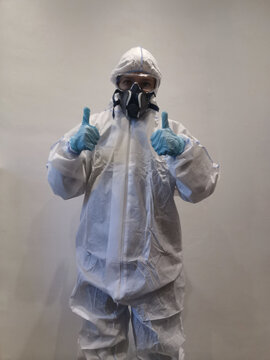 A person wearing full-body personal protective equipment (PPE), including a white hazmat suit, mask, and blue gloves, giving a thumbs up