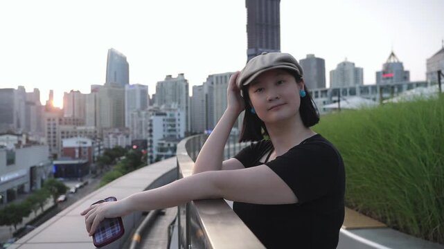 A woman in a light beret and black top poses on a curved rooftop with a city skyline at dusk in the background.