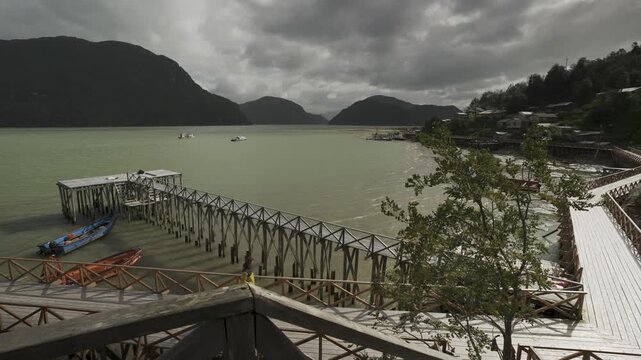 Tourist walking along the traditional wooden walkways of Caleta Tortel, featuring scenic piers, moored boats, and a mountainous fjord landscape under a cloudy sky in Patagonia, Chile.