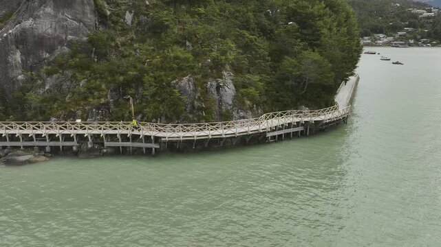Close-up aerial view of a person walking along the iconic wooden footbridge built on stilts along the rugged cliffside of Caleta Tortel, Patagonia, Chile.