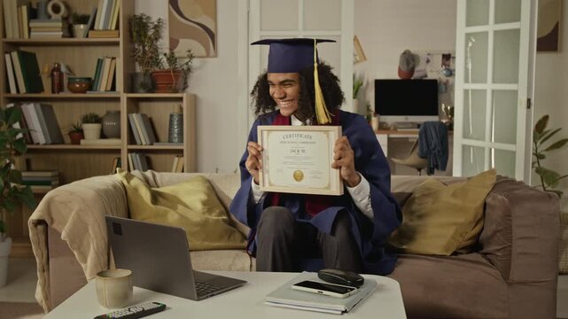Medium long shot of African American teenager wearing graduation gown sitting on sofa, showing certificate to laptop camera. Ideal for online education and remote learning advertisements